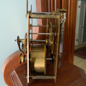 May include: Close-up of a brass clock mechanism with gears and a winding key. The mechanism is mounted on a wooden surface.