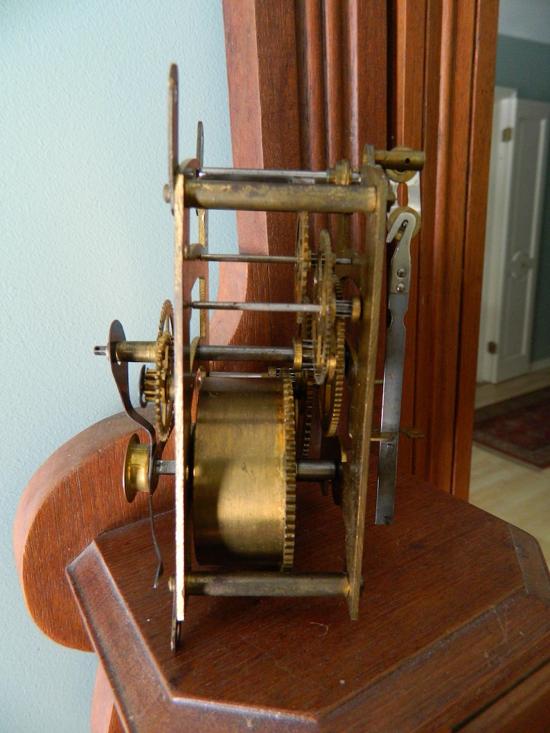 May include: Close-up of a brass clock mechanism with gears and a winding key. The mechanism is mounted on a wooden surface.