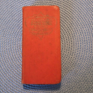 May include: A vintage, rectangular, red book with gold lettering. The book's cover features the title "Abridged Standard Dictionary of the English Language" in an ornate, framed design. The book is set against a textured, blue background.