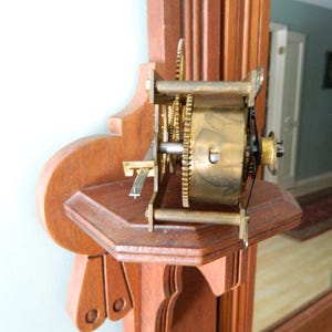 May include: Close-up of a wooden clock mechanism with brass gears and a metal rod. The mechanism is mounted on a wooden shelf with intricate carvings.