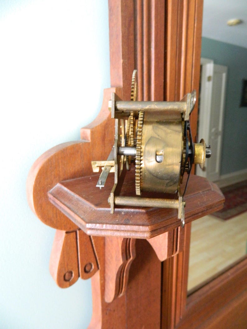 May include: Close-up of a wooden clock mechanism with brass gears and a metal rod. The mechanism is mounted on a wooden shelf with intricate carvings.