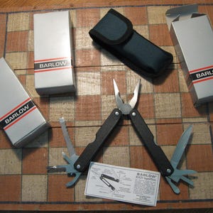 May include: A multi-tool with black handles and various folding tools, including pliers and a knife, is displayed open. The tool is accompanied by a black carrying case and several silver boxes with the word "BARLOW" printed on them.