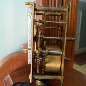 May include: Close-up of a brass clock mechanism with gears and a winding key. The mechanism is mounted on a wooden surface.