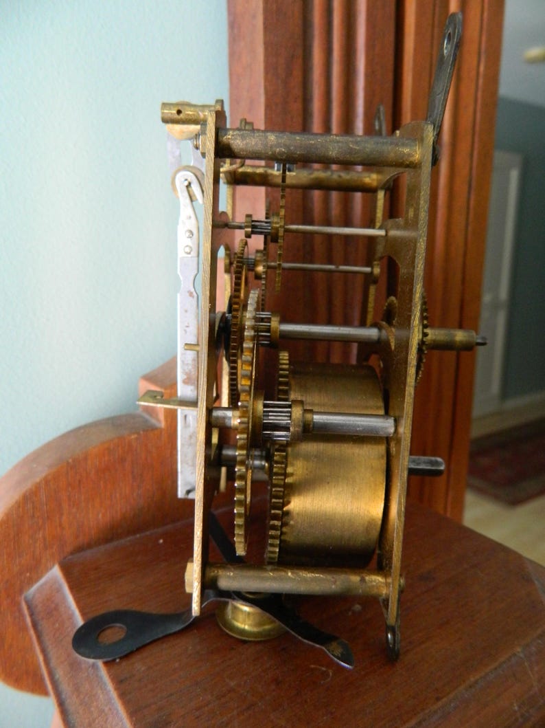 May include: Close-up of a brass clock mechanism with gears and a winding key. The mechanism is mounted on a wooden surface.