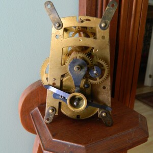 May include: Close-up of a brass clock mechanism with intricate gears and levers. The mechanism is mounted on a wooden shelf.