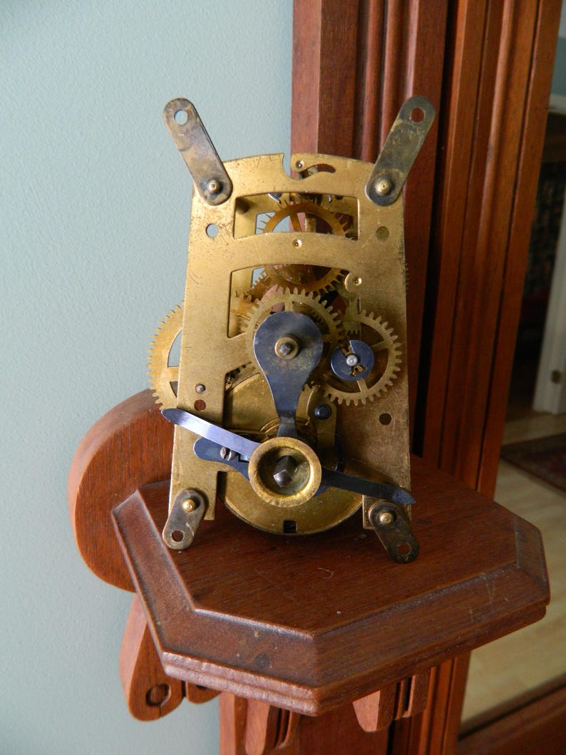 May include: Close-up of a brass clock mechanism with intricate gears and levers. The mechanism is mounted on a wooden shelf.