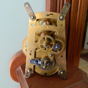 May include: Close-up of a brass clock mechanism with intricate gears and a winding key. The mechanism is mounted on a wooden base.