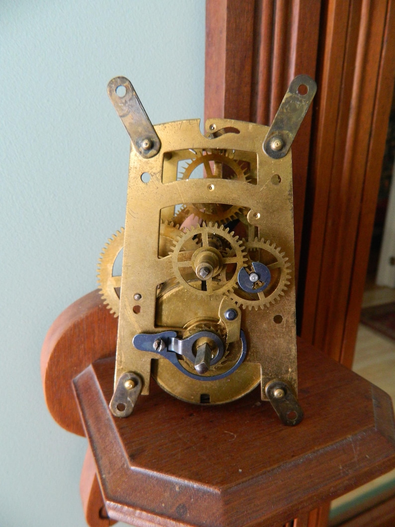 May include: Close-up of a brass clock mechanism with intricate gears and a winding key. The mechanism is mounted on a wooden base.