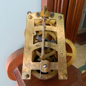 May include: Close-up of a gold-toned clock mechanism with gears and a winding key. The mechanism is mounted on a wooden surface.