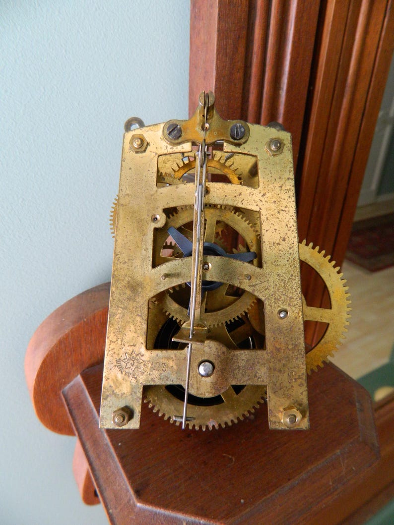 May include: Close-up of a gold-toned clock mechanism with gears and a winding key. The mechanism is mounted on a wooden surface.