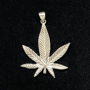 May include: A silver pendant in the shape of a marijuana leaf. The leaf is detailed with veins and has a smooth, polished finish.