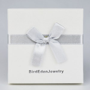 May include: A square, off-white jewelry box with a silver ribbon and bow. The box has a textured surface and the text "BirdEdenJewelry" printed in gray on the front.