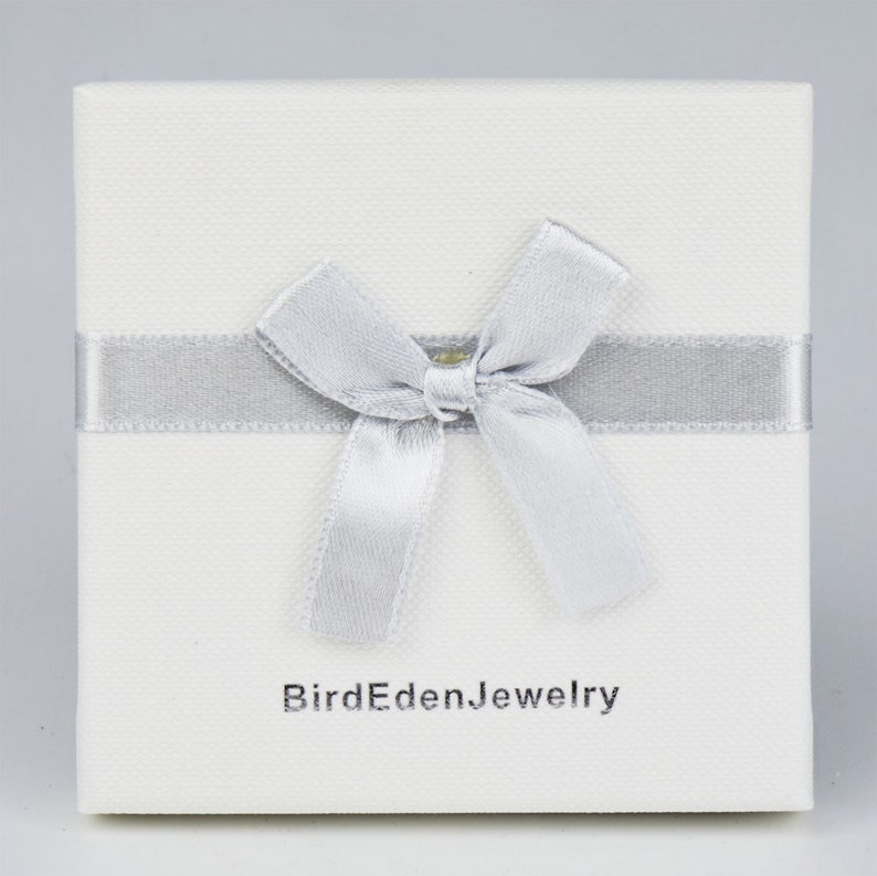 May include: A square, off-white jewelry box with a silver ribbon and bow. The box has a textured surface and the text "BirdEdenJewelry" printed in gray on the front.