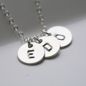 Peut inclure: Collier en argent avec trois breloques rondes, chacune avec une lettre estampée. Les lettres sont E, D et O.