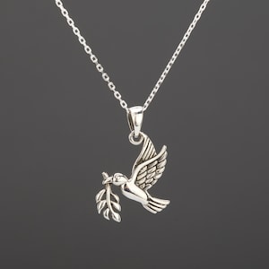 May include: A silver-toned pendant necklace featuring a dove carrying an olive branch. The dove is in mid-flight, with detailed wing and feather designs. The necklace chain is delicate and silver, set against a dark background.