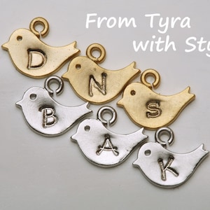 May include: A collection of bird-shaped charms in gold and silver tones, each with a different letter engraved on its body. The charms are arranged diagonally, with the text "From Tyra with Style" in the upper right corner.