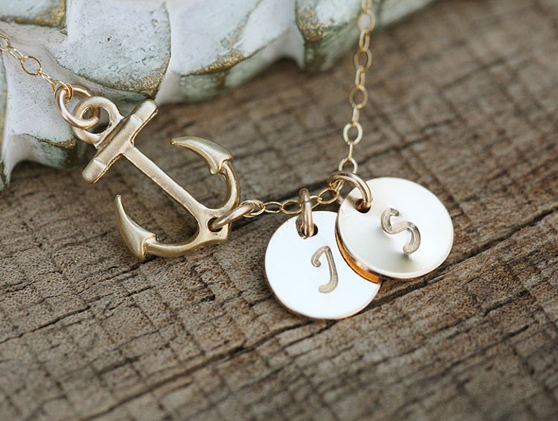 May include: A gold-colored necklace featuring an anchor pendant and two circular charms. The charms are engraved with the letters "J" and "S". The necklace is displayed on a wooden surface.