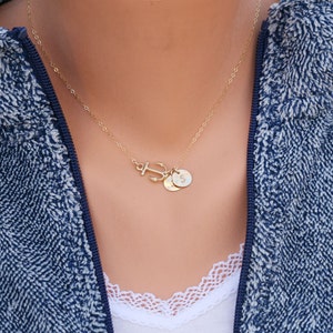 May include: A gold-colored necklace featuring an anchor pendant and two small circular charms. The necklace is worn against a blue and white textured jacket. The charms appear to have initials on them.