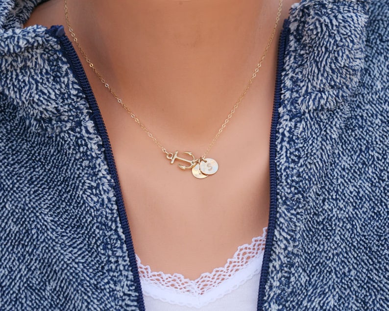May include: A gold-colored necklace featuring an anchor pendant and two small circular charms. The necklace is worn against a blue and white textured jacket. The charms appear to have initials on them.