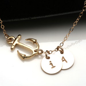 May include: Gold-colored necklace featuring an anchor pendant and two small, round charms. The charms are engraved with the letters "i" and "A". The necklace chain is delicate and the anchor is a nautical design.