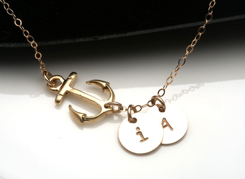 May include: Gold-colored necklace featuring an anchor pendant and two small, round charms. The charms are engraved with the letters "i" and "A". The necklace chain is delicate and the anchor is a nautical design.