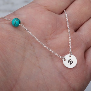 May include: A silver necklace with a turquoise bead and a silver charm with the letter "E" engraved on it.