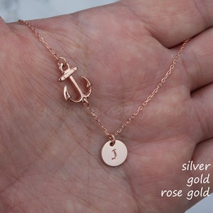 May include: Rose gold necklace featuring an anchor charm and a small circular pendant with the letter "J". The necklace is displayed on a hand, showcasing its delicate chain and design. The text "silver gold rose gold" is visible.