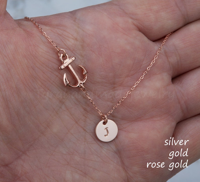 May include: Rose gold necklace featuring an anchor charm and a small circular pendant with the letter "J". The necklace is displayed on a hand, showcasing its delicate chain and design. The text "silver gold rose gold" is visible.