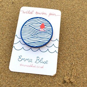 May include: A white card with blue and pink text that reads "wild swim pin" and "Emma Blue emmablue.co.uk". A blue and white circle with a pink figure swimming in waves is attached to the card.
