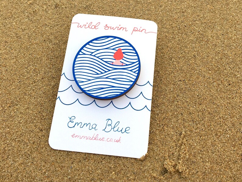 May include: A white card with blue and pink text that reads "wild swim pin" and "Emma Blue emmablue.co.uk". A blue and white circle with a pink figure swimming in waves is attached to the card.