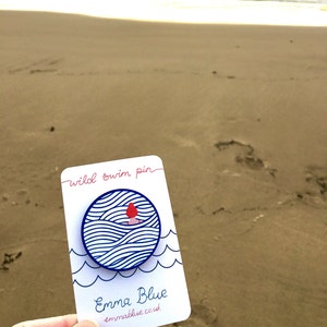 May include: A white card with blue and red details. The card says "wild swim pin" and "Emma Blue emmablue.co.uk". The card features a blue circle with a white background and a red figure swimming in blue waves.