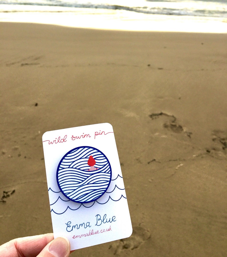 May include: A white card with blue and red details. The card says "wild swim pin" and "Emma Blue emmablue.co.uk". The card features a blue circle with a white background and a red figure swimming in blue waves.