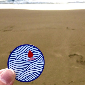 May include: A blue and white sticker with a stylised illustration of a person with red hair swimming in waves.