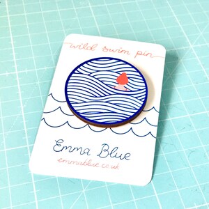 May include: A wooden pin featuring a blue and white wave design with a small red figure swimming in the waves. The pin is displayed on a white card with the text "wild swim pin" and "Emma Blue" with the website address "emmablue.co.uk".