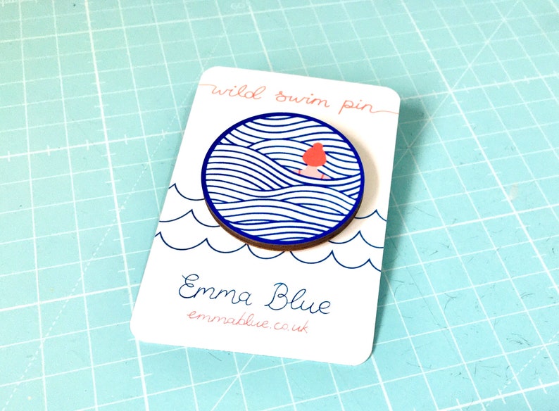 May include: A wooden pin featuring a blue and white wave design with a small red figure swimming in the waves. The pin is displayed on a white card with the text "wild swim pin" and "Emma Blue" with the website address "emmablue.co.uk".