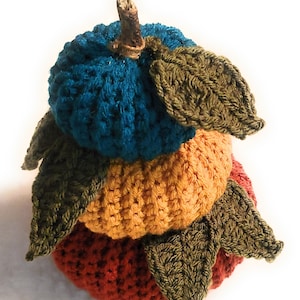 May include: A stack of three crocheted pumpkins in teal, orange, and red with green crocheted leaves.