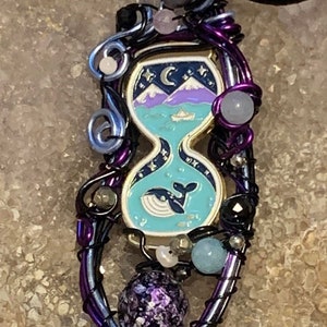 May include: A purple and silver wire-wrapped pendant featuring a glass hourglass charm with a night sky and a whale design. The pendant is adorned with various beads, including a purple iridescent bead.