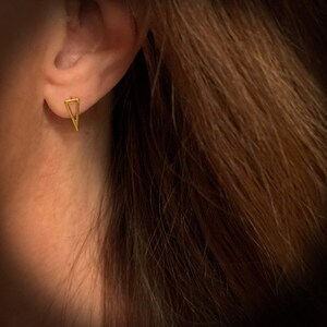 May include: A small gold-toned stud earring shaped like an open triangle. The earring is positioned in the earlobe, featuring a clean, geometric outline with a pointed bottom and a flat top edge, showcasing a polished metallic finish throughout.