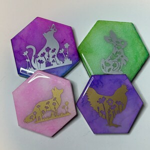 May include: Four hexagonal coasters featuring different animal silhouettes and floral designs. The coasters are made of resin and have a watercolour effect. The animals are a cat, a rabbit, a fox, and a chicken. The colours are purple, green, pink, and blue.