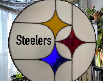 May include: A round stained glass window with a black metal frame. The window features a stylized star design with yellow, red, and blue glass pieces. The word "Steelers" is written in black lettering on the white glass.