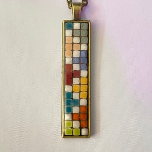May include: A long, rectangular pendant necklace with a mosaic design. The mosaic is made up of small, colorful squares in shades of blue, green, yellow, orange, red, white, and brown. The pendant is attached to a bronze chain.