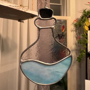 May include: A stained glass hanging ornament shaped like a bottle with a blue liquid inside. The bottle is made of clear glass with a silver metal frame. The ornament is hanging from a silver chain.