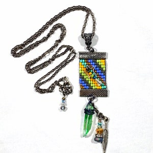 Op de afbeelding: Een kralenketting met een kleurrijke rechthoekige hanger met een stervormig ontwerp. De hanger is bevestigd aan een zilveren ketting met een groene kristal en een veerbedel.