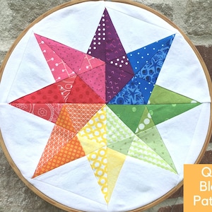 May include: A colourful quilt block pattern featuring a star shape with a rainbow of colours, including red, orange, yellow, green, blue, purple, and pink. The star is made up of smaller triangles, each with a different pattern. The quilt block is framed in a wooden hoop.
