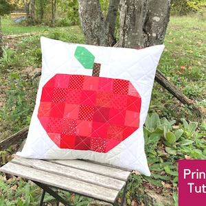 Könnte beinhalten: Ein rotes und weißes Patchwork-Apfelkissen mit grünem Stiel und Blatt. Das Kissen liegt auf einer Holzbank in einem grasbewachsenen Bereich. Der Text "Druckbarer Leitfaden" ist in der unteren rechten Ecke des Bildes sichtbar.