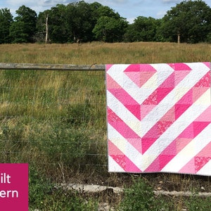 May include: A pink and white quilt pattern with a chevron design. The quilt is hanging on a wooden fence against a backdrop of a grassy field and trees.