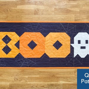 Pode incluir: Um padrão de acolchoado para um corredor de mesa de Halloween com a palavra "BOO" em tecido laranja e preto com um fantasma branco no centro. O fundo é um tecido azul escuro com um design de teia de aranha.