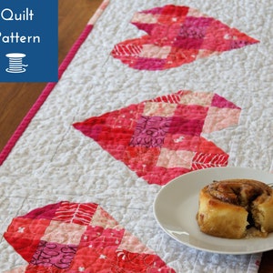 Può includere: Un runner da tavolo bianco con tre cuori in patchwork rosa e rosso. Il runner è trapuntato e ha uno sfondo bianco. Un piatto con un dolce alla cannella è sul runner da tavolo.
