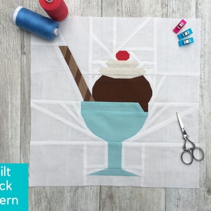 Puede incluir: Un bloque de tela para acolchar con un helado de dibujos animados en un vaso azul. El helado tiene helado de chocolate, crema batida y una cereza. Las palabras "Quilt Block Pattern" están en un cuadro azul. Suministros de costura en el fondo.