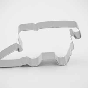 May include: A silver-colored cookie cutter in the shape of a combine harvester. The cutter is made of metal and has a simple, clean design. The combine harvester cookie cutter is a great addition to any baking collection.